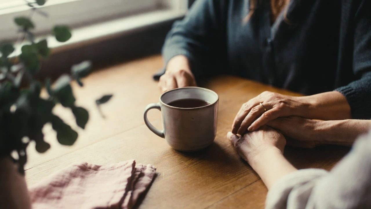 A nurse holding a client's hand across a kitchen table beside a mug of tea
